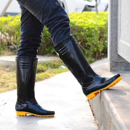 Homme portant des bottes noires hautes avec une semelle jaune. Il a un pied posé sur un rebord et on voit derrière un jardin.