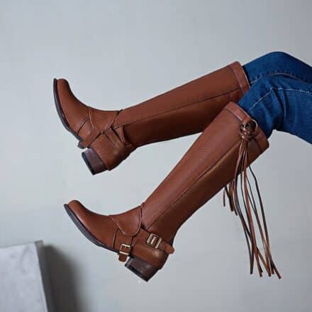 Photo d'une paire de bottes cavalière marron à franges. Les bottes sont portées avec un jean sur un fond gris avec une plaque en marbre en bas à gauche.