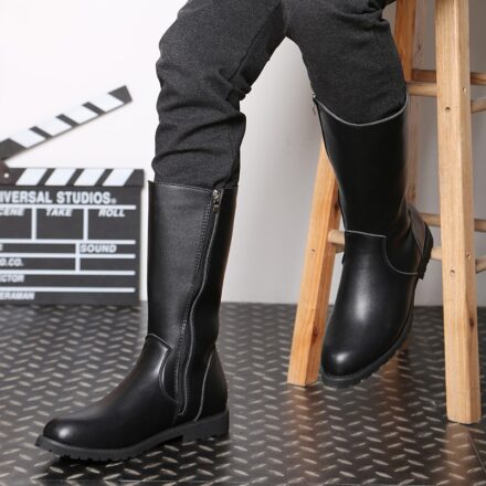 Bottes noires portées par un homme en pantalon gris assis sur un tabouret en bois sur un fond blanc et un salle noir. En arrière plan se trouve un calp de cinéma en décoration