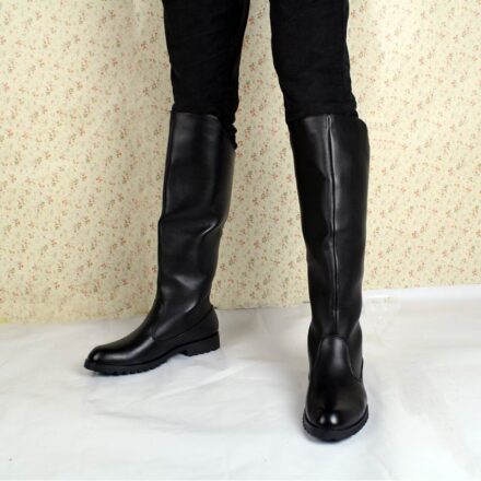 Jambes d'un hommes portant des bottes hautes en cuir noir sur un fond beige et un sol blanc