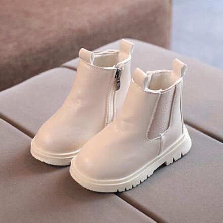 On voit de très jolies petites bottines couleur crème pour petite fille. Elles sont posées sur un canapé gris.