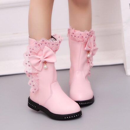 On voit les jambes et les pieds s'une petite fille qui porte des bottes roses avec un noeud, de la dentelle et des strass. La semelle est noire et le sol est gris.