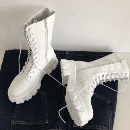 Bottes mi-hautes blanches à lacets pour femme. Bottes à plateforme et zip sur le côté pour les enfiler. Posées sur une commode noire, une botte est tombée sur le côté.
