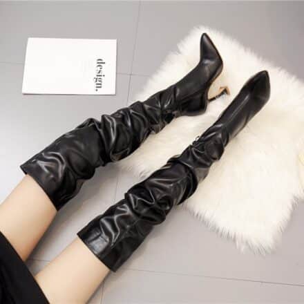 Bottes noires cuiisardes à talon sur tapis en fourrure blanc sur un sol gris avec un livre et un chapeau