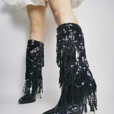 jeune femme qui porte une jupe blanche courte et dont on ne voit que les jambes, porte une paire de botte vintage noires, à talons aiguilles, avec des franges en strass