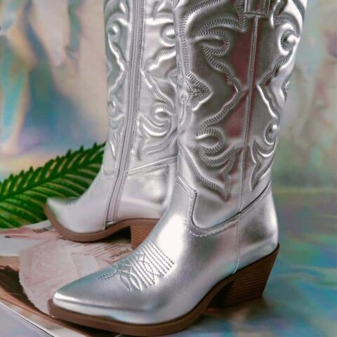 Paire de bottes style cowboy grise effet métallisé, avec semelle et talon marron. De trois-quarts les deux. Botte droite vue de l'intérieur avec la fermeture éclair, botte de gauche vue de l'extérieur. Sur fond papier argenté.