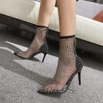 Mannequin femme portant des bottines pointues en maille scintillantes, on ne voit que ses pieds et un bout de ses mollets sur une moquette grise et on voit un canapé beige clair en arrière-plan.
