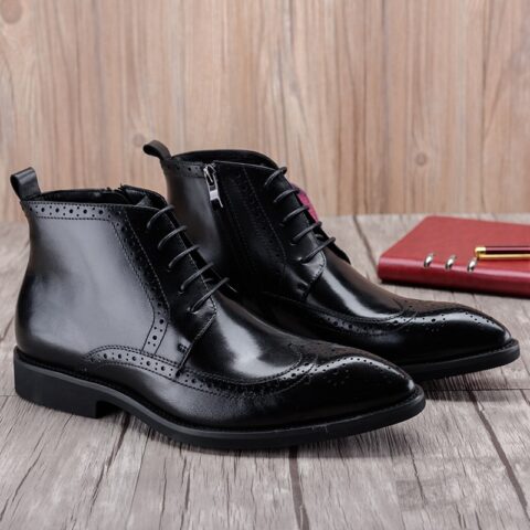 Paire de bottine noires homme, style derby à lacets et fermeture éclair latéral. Sur un sol en parquet gris, on voit un mur en bois en fond et un calepin rouge sur la droite.
