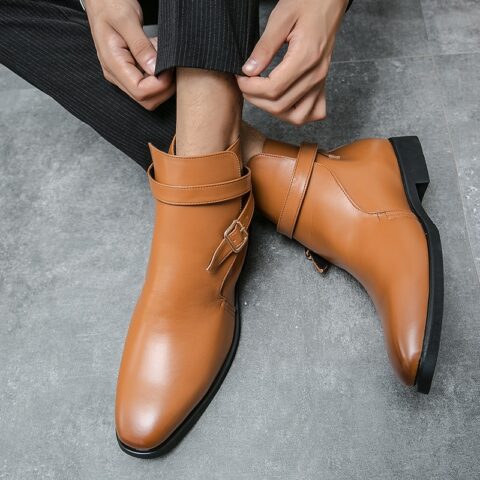 Mannequin homme portant des bottes marron clair avec sangle à boucle au niveau de la cheville. On ne voit que ses pieds et ses chevilles, ainsi que ses deux mains qui ajustent son bas de pantalon. Il a les jambes qui se croisent au niveau des chevilles avec un pied à plat au sol et l'autre posé de profil.
